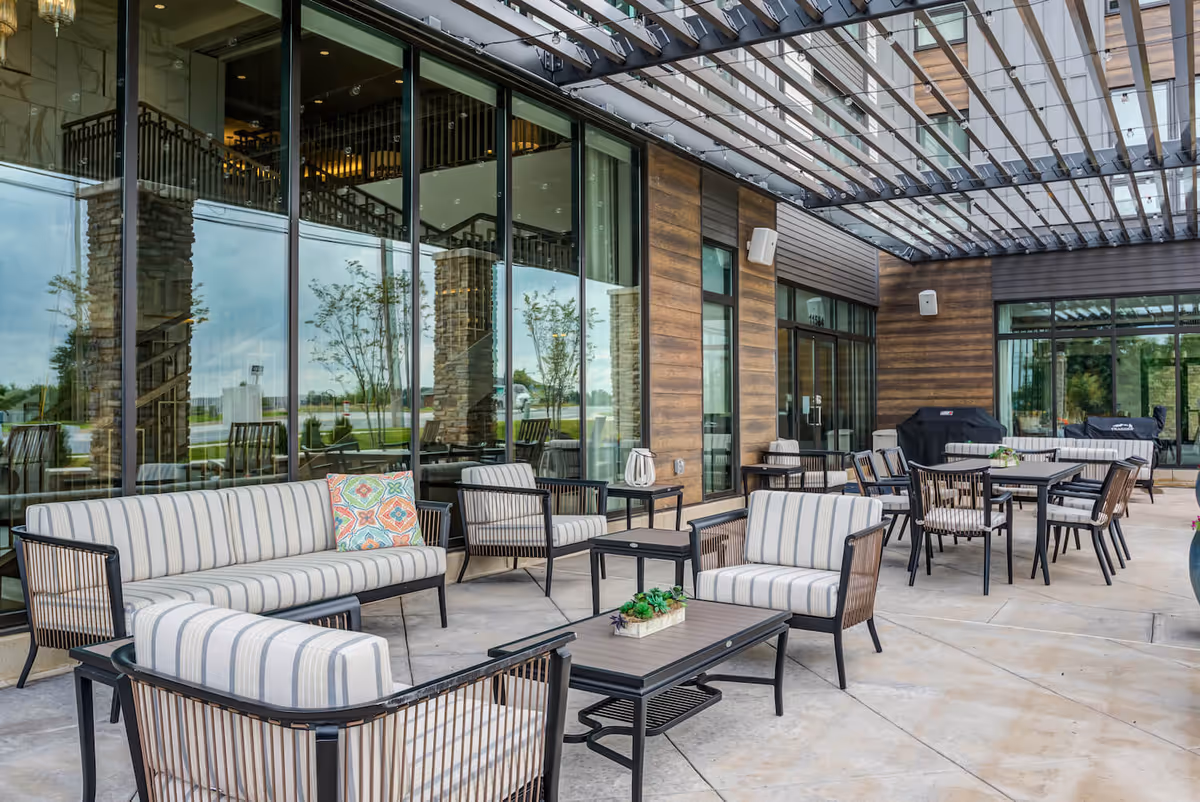Outdoor patio area at Arbor Terrace Fulton featuring striped cushioned seating including sofas and armchairs, several tables, a grill, and a pergola overhead with string lights. The patio is adjacent to a building with large glass windows and wood-paneled walls.
