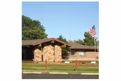 Single-story building with a stone entrance and an American flag on a flagpole in front, surrounded by green grass and trees under a clear blue sky.
