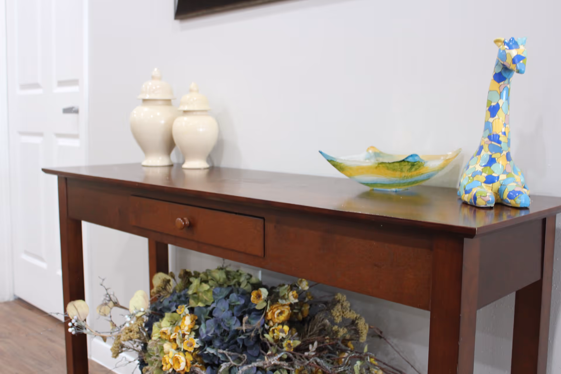 A wooden console table against a light-colored wall with decorative items on top including two cream-colored ceramic jars, a colorful glass bowl, and a blue and yellow mosaic giraffe figurine. Below the table is a floral arrangement with yellow and blue flowers. A white door is visible in the background.