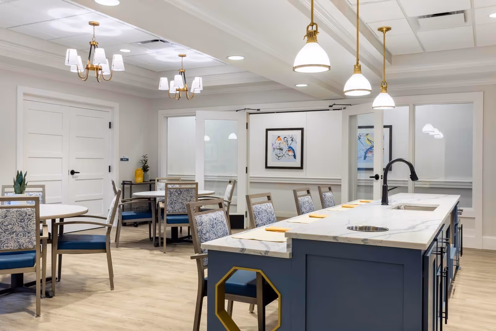A bright and modern dining room in a senior living facility featuring round tables with patterned chairs, a large kitchen island with a marble countertop and sink, pendant lights hanging above the island, and framed bird artwork on the walls.