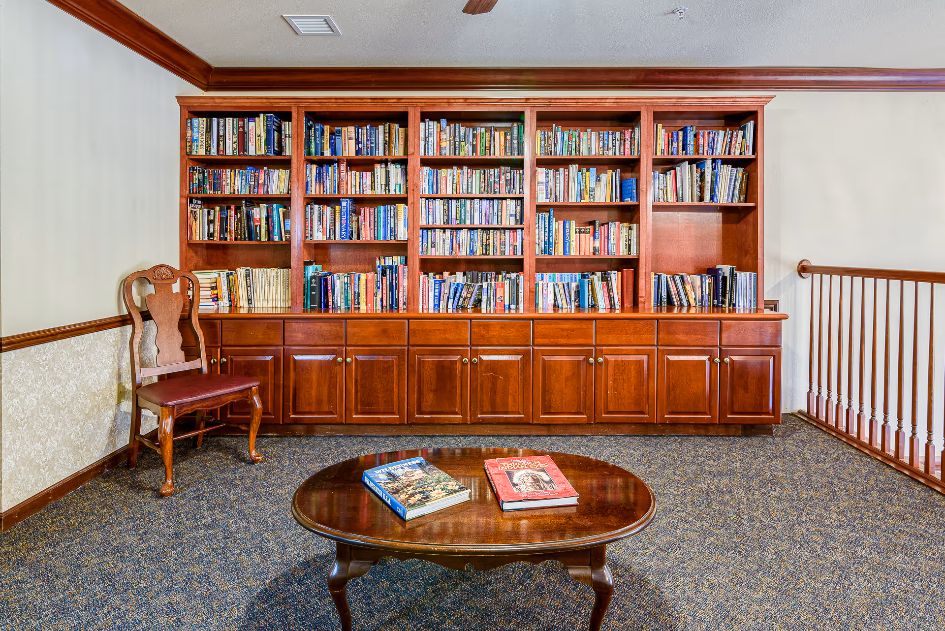 Interior view of a cozy reading area with a large wooden bookshelf filled with books, a wooden chair with a cushioned seat to the left, and a wooden coffee table with two books on it in the foreground. The room has carpeted flooring and a wooden railing on the right side.