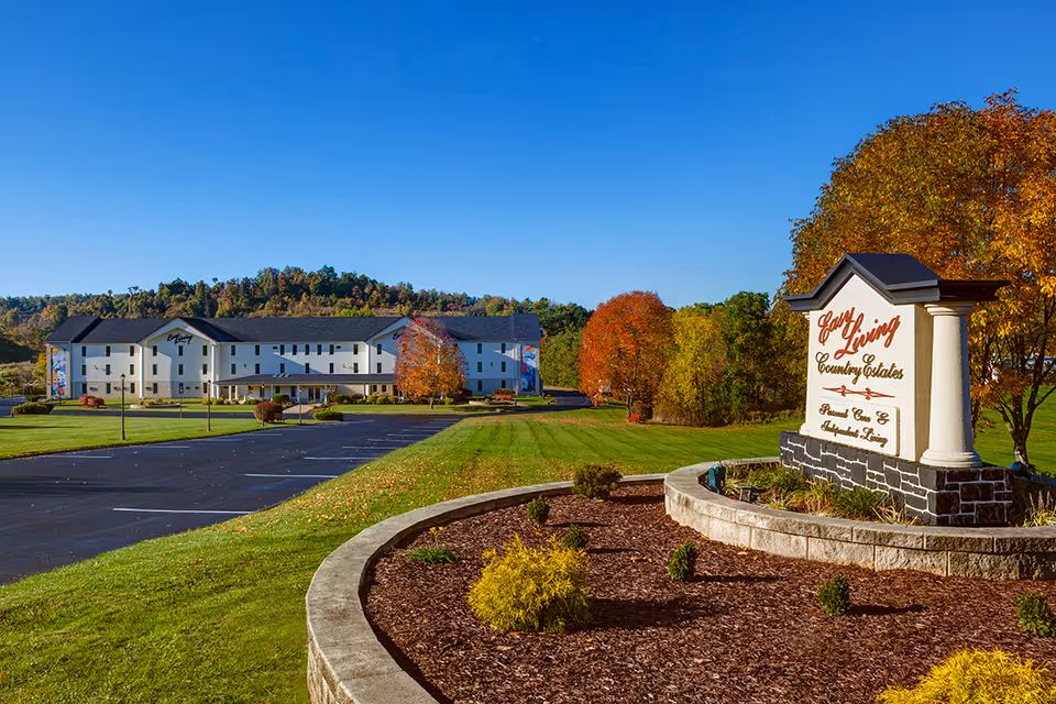 Wide exterior view of Easy Living Country Estates facility on a clear sunny day with a large parking lot, manicured lawn, autumn trees, and a prominent sign in the foreground displaying the facility name.