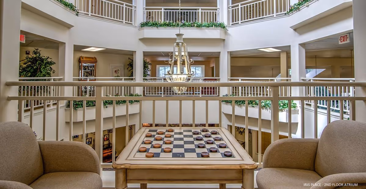 Indoor atrium area with a checkerboard table set between two beige armchairs. The space features white railings, green plants along the upper balcony edges, and a hanging chandelier light fixture. The atrium is open to multiple floors with visible windows and exit signs.