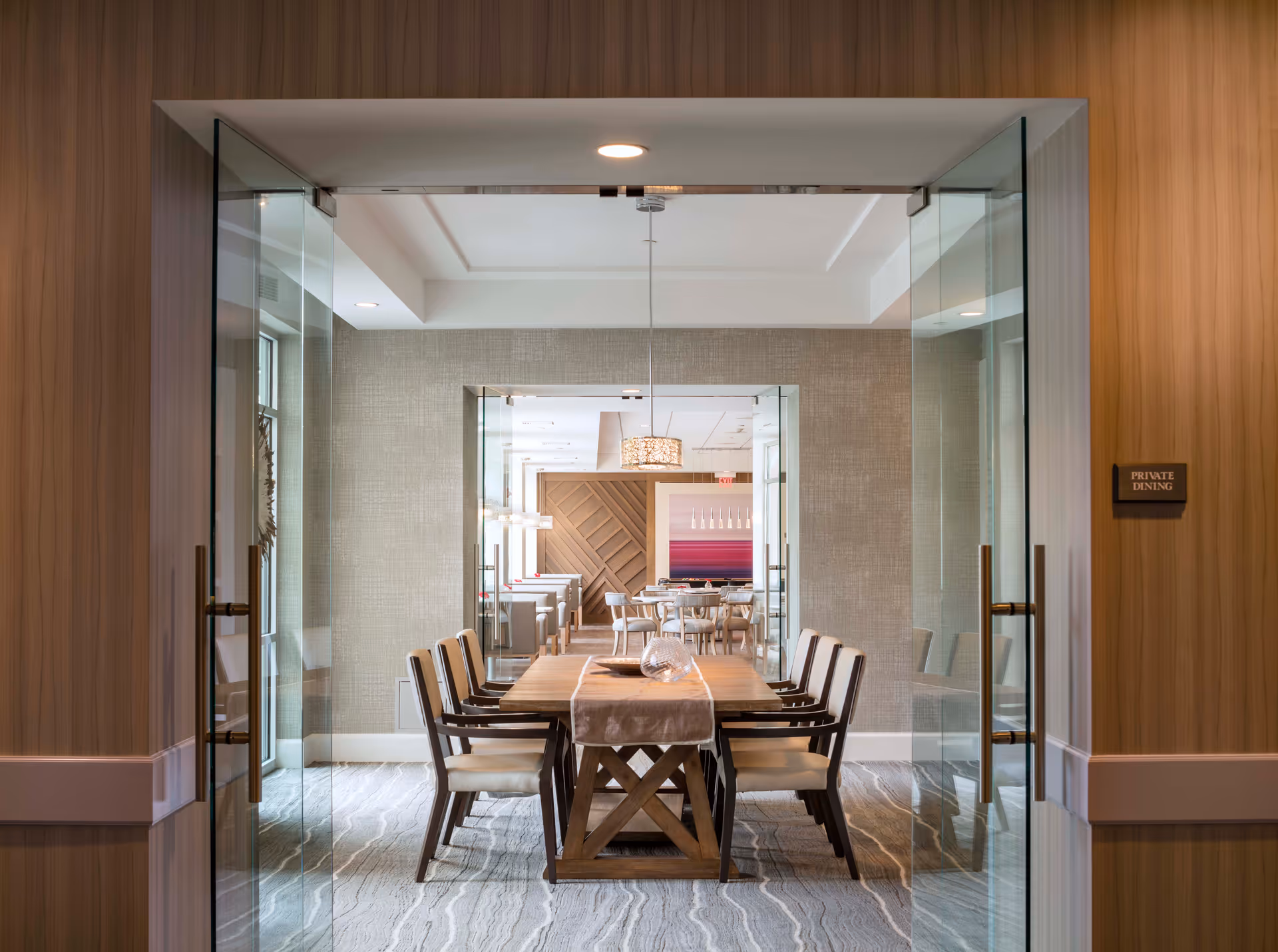 View through glass double doors into a private dining room with a long wooden table and eight cushioned chairs. The room has a modern design with neutral tones, textured walls, and a decorative light fixture hanging above the table. A sign on the right wall reads 'PRIVATE DINING'.