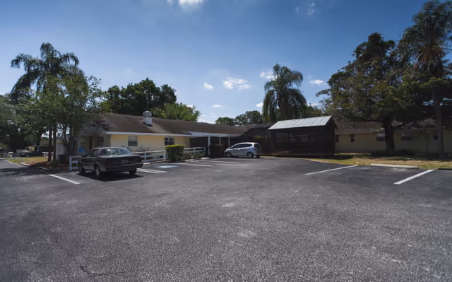 Parking lot in front of a single-story building with a screened porch area, surrounded by trees and clear blue sky.