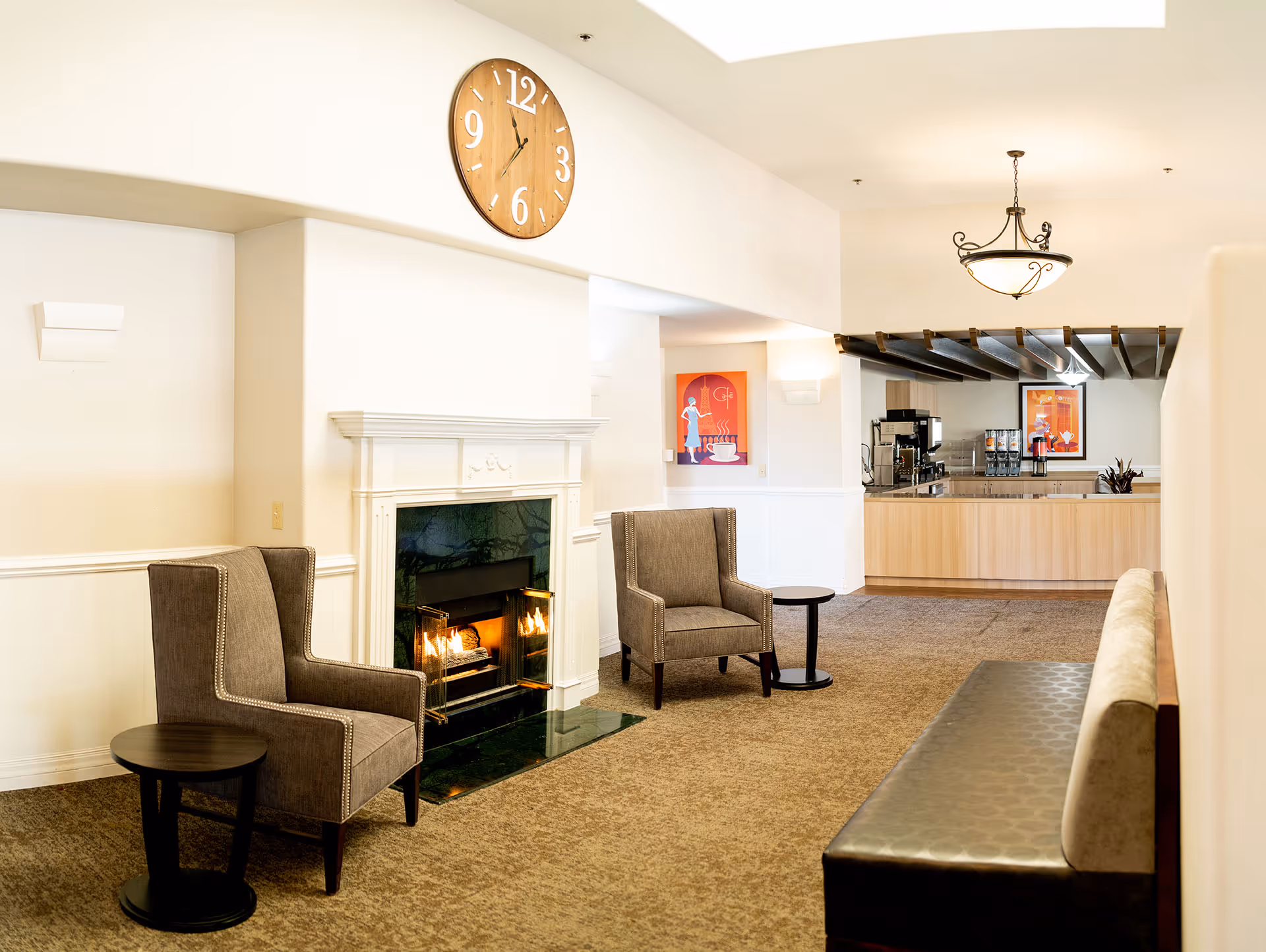 A cozy senior living common area with two gray armchairs and a small round table in front of a lit fireplace with a white mantel. Above the fireplace is a large wooden clock showing the time as 11:55. To the right, there is a coffee station with a counter, coffee machines, and colorful artwork on the walls. A cushioned bench is visible on the right side of the image.