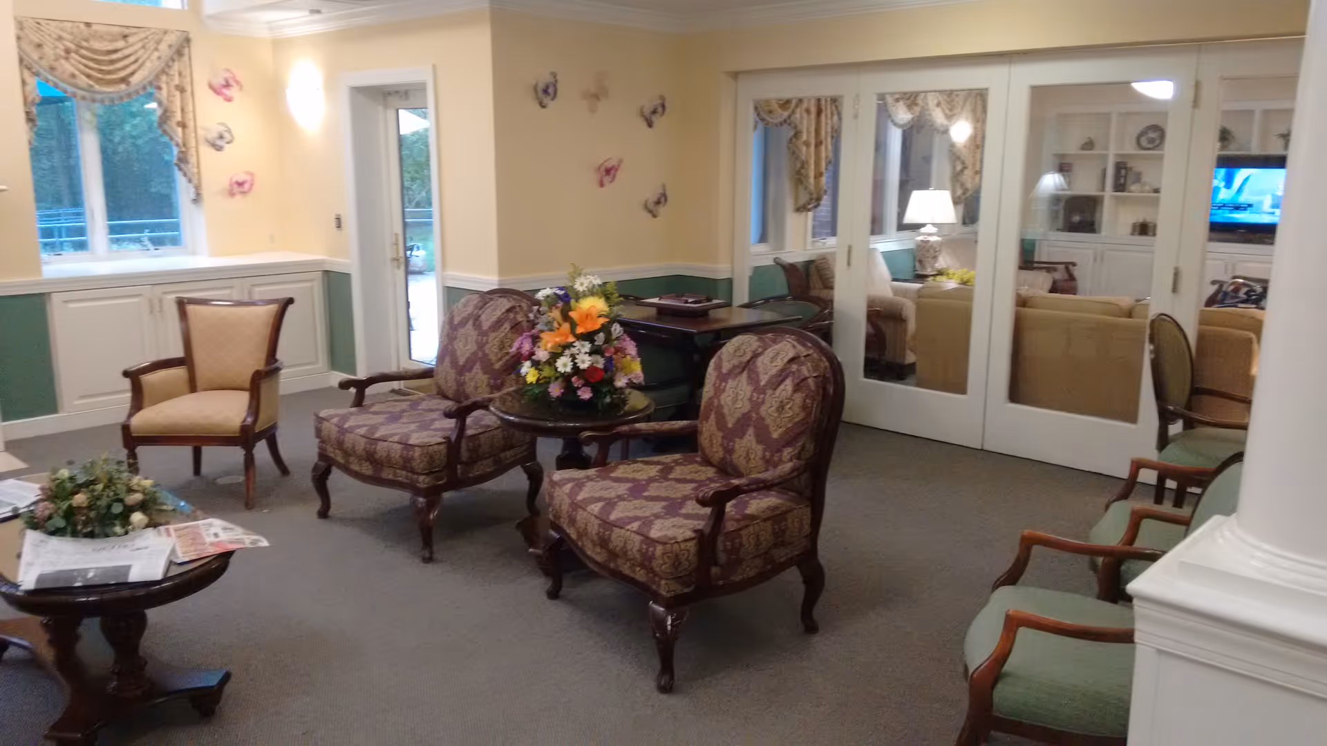 A cozy sitting area in a senior living facility with several upholstered armchairs arranged around small wooden tables. One table has a colorful flower arrangement, and another has newspapers and a floral centerpiece. The room features beige walls with green wainscoting, a window with decorative curtains, and glass sliding doors leading to another room with more seating and a television.