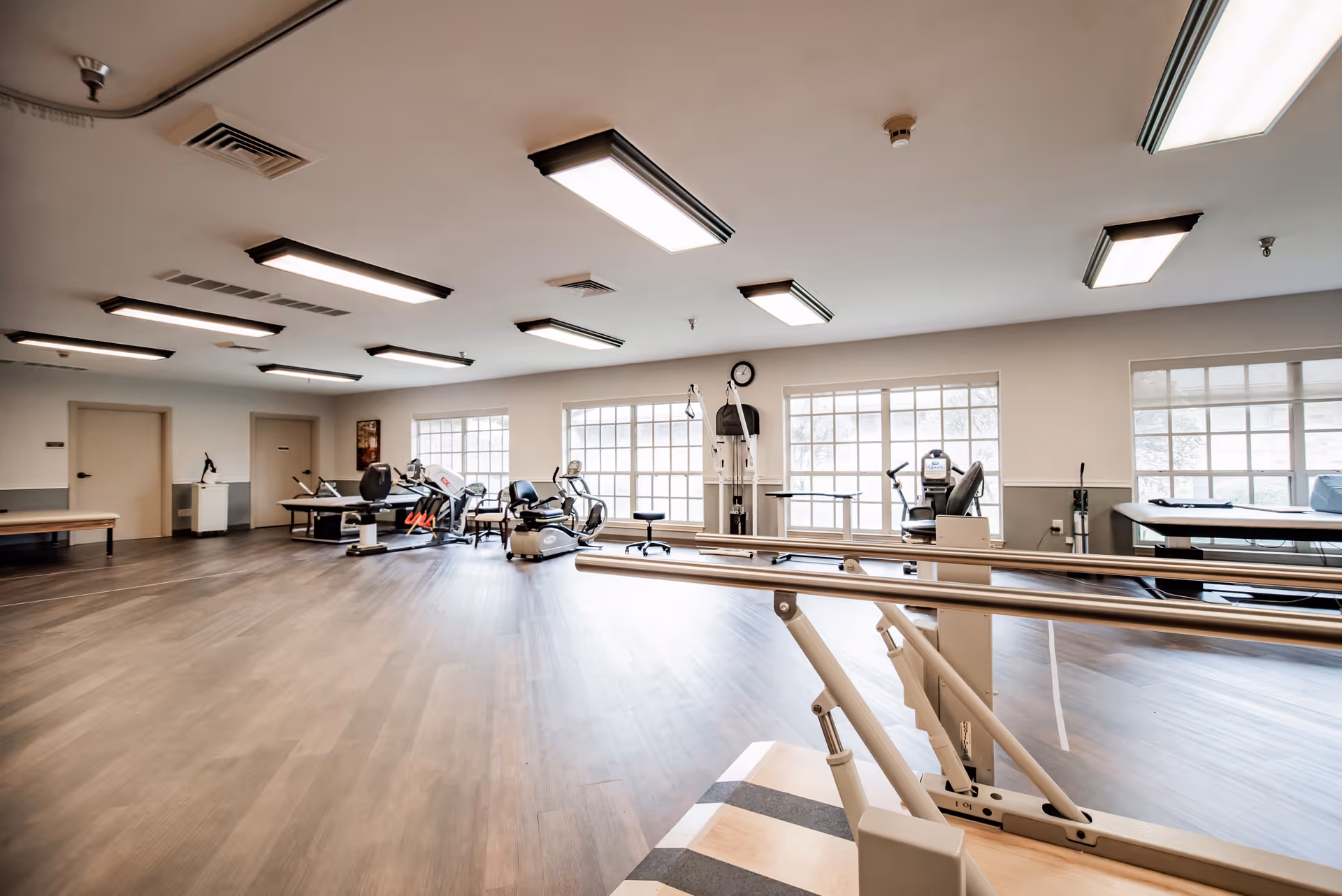 A spacious rehabilitation room with wooden flooring and large windows letting in natural light. The room contains various exercise and physical therapy equipment including stationary bikes, parallel bars for walking exercises, and other rehabilitation machines. The ceiling has multiple rectangular fluorescent lights, and there are a few closed doors along the left wall.