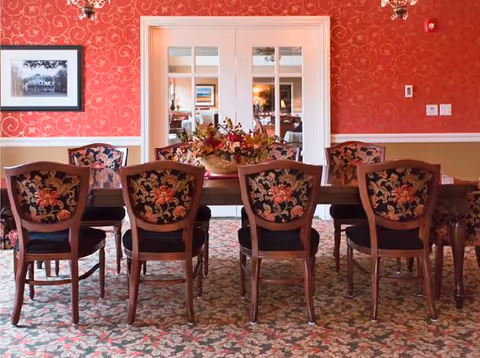 A formal dining room with a long wooden table, floral-upholstered chairs, a centerpiece, and red patterned wallpaper.