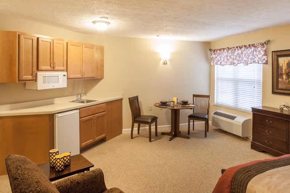 A cozy studio apartment interior featuring a small kitchenette with wooden cabinets, a microwave, a sink, and a mini fridge. There is a round dining table set for two with dark wooden chairs near a window with blinds and a floral valance. The room also includes a patterned armchair, a wooden dresser, and part of a bed with a brown and red comforter. The walls are painted beige and the floor is carpeted.
