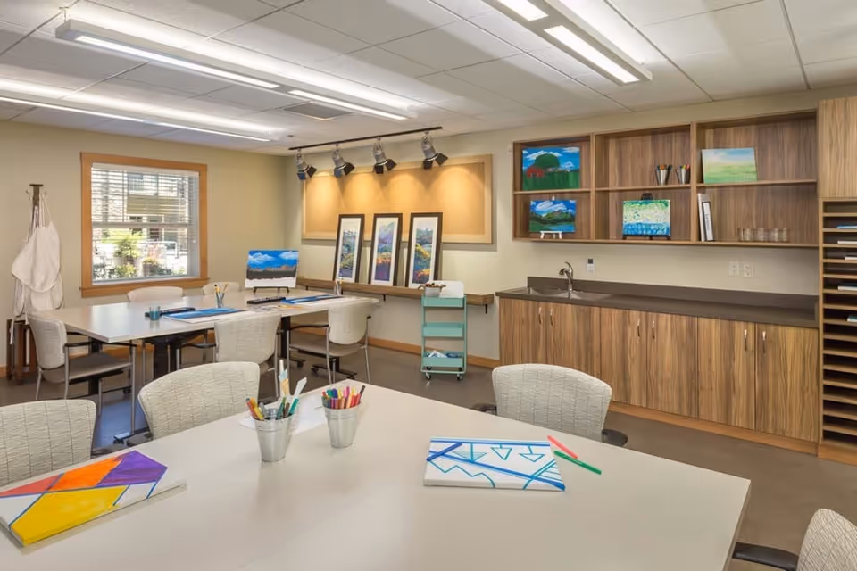 Bright activity room with multiple tables and chairs, art supplies and canvases on the tables, framed paintings on a wall, and wood cabinets with a sink.