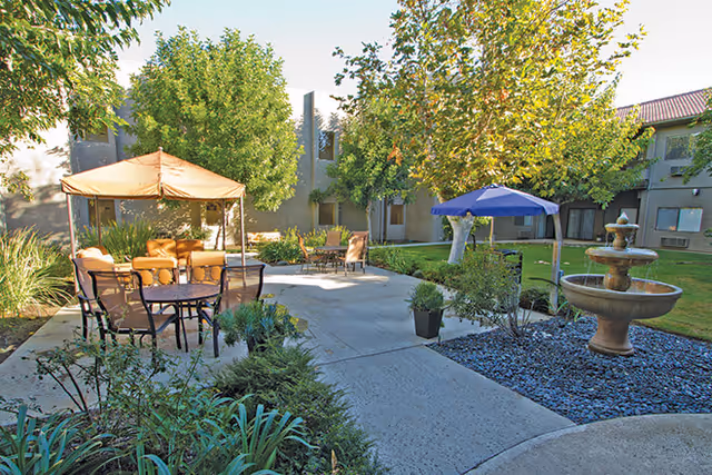 Outdoor courtyard area at Brookdale Chatsworth featuring a paved walkway, patio tables with umbrellas, chairs, greenery, and a multi-tiered water fountain surrounded by rocks.