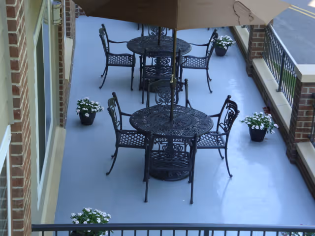 Outdoor patio area with black wrought iron tables and chairs, each table shaded by a large umbrella. Several potted white flowering plants are placed along the edges of the patio, which is bordered by brick walls and a metal railing.