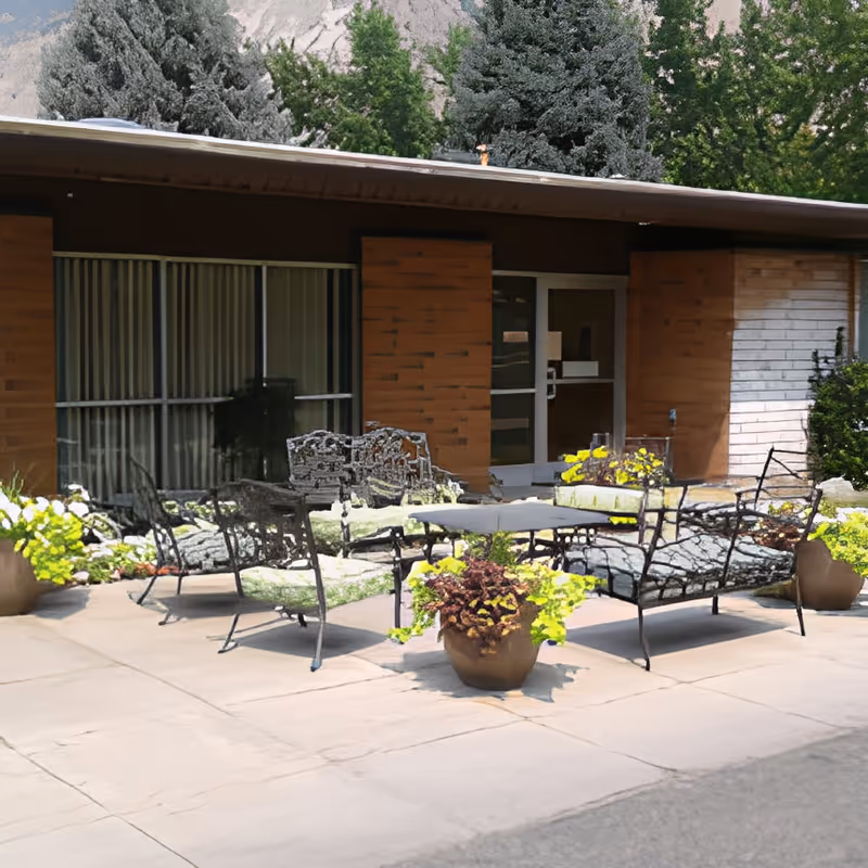 Outdoor patio area with metal chairs and tables surrounded by potted plants and greenery, adjacent to a building with large windows and a door.
