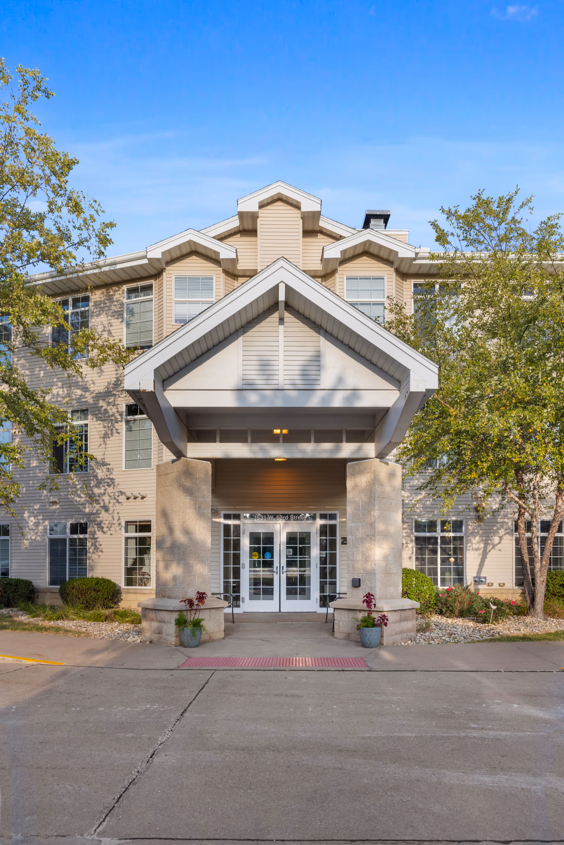 Front exterior view of a senior living facility building with a covered entrance supported by two stone pillars, glass double doors, and surrounding trees and shrubs under a clear blue sky.