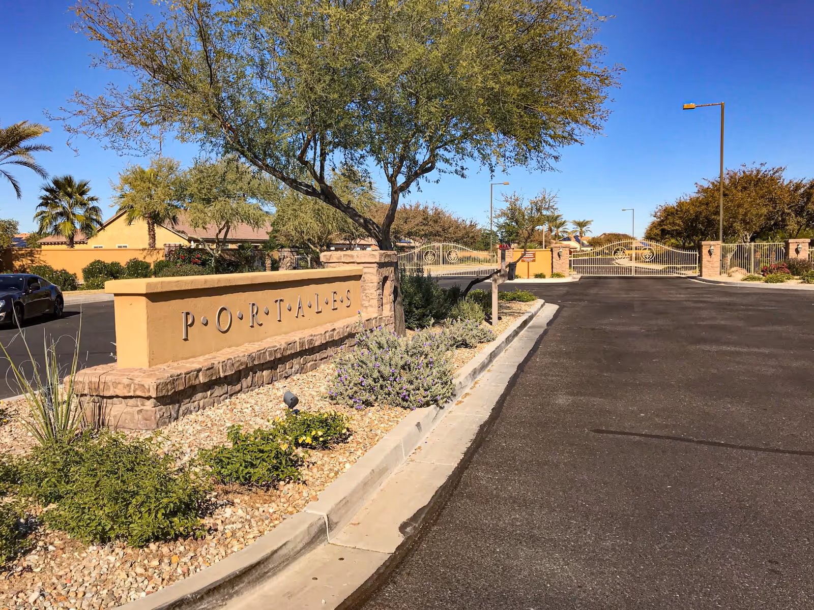 Gated community entrance with a stone sign reading 'PORTALES', landscaped rock beds, and a paved driveway under a clear blue sky.