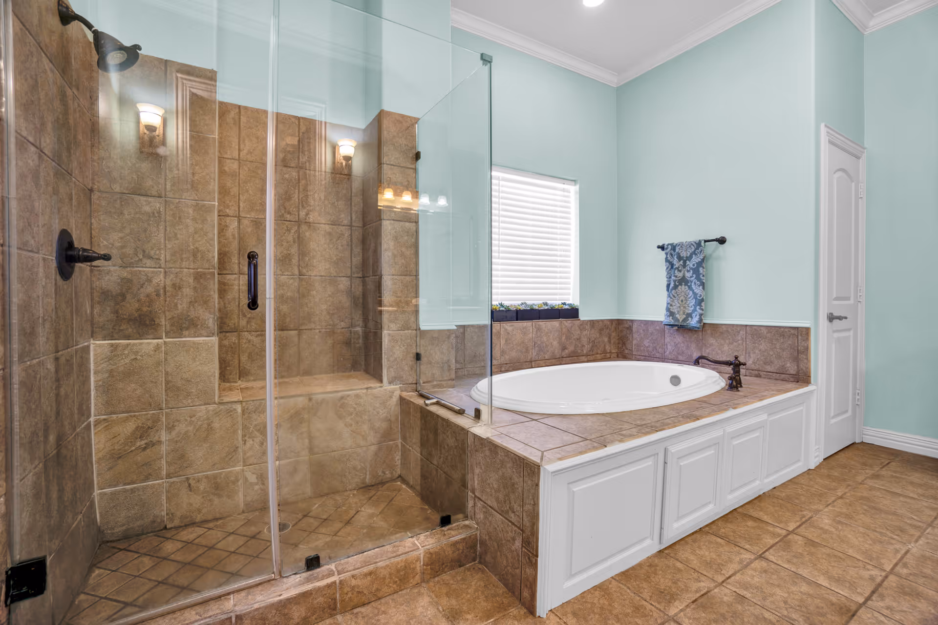 A bathroom featuring a glass-enclosed shower with brown tiles and a built-in bench, next to a large white bathtub surrounded by matching brown tiles. The walls are painted light blue, and there is a window with white blinds above the bathtub. A blue patterned towel hangs on a black towel rack next to a white door.