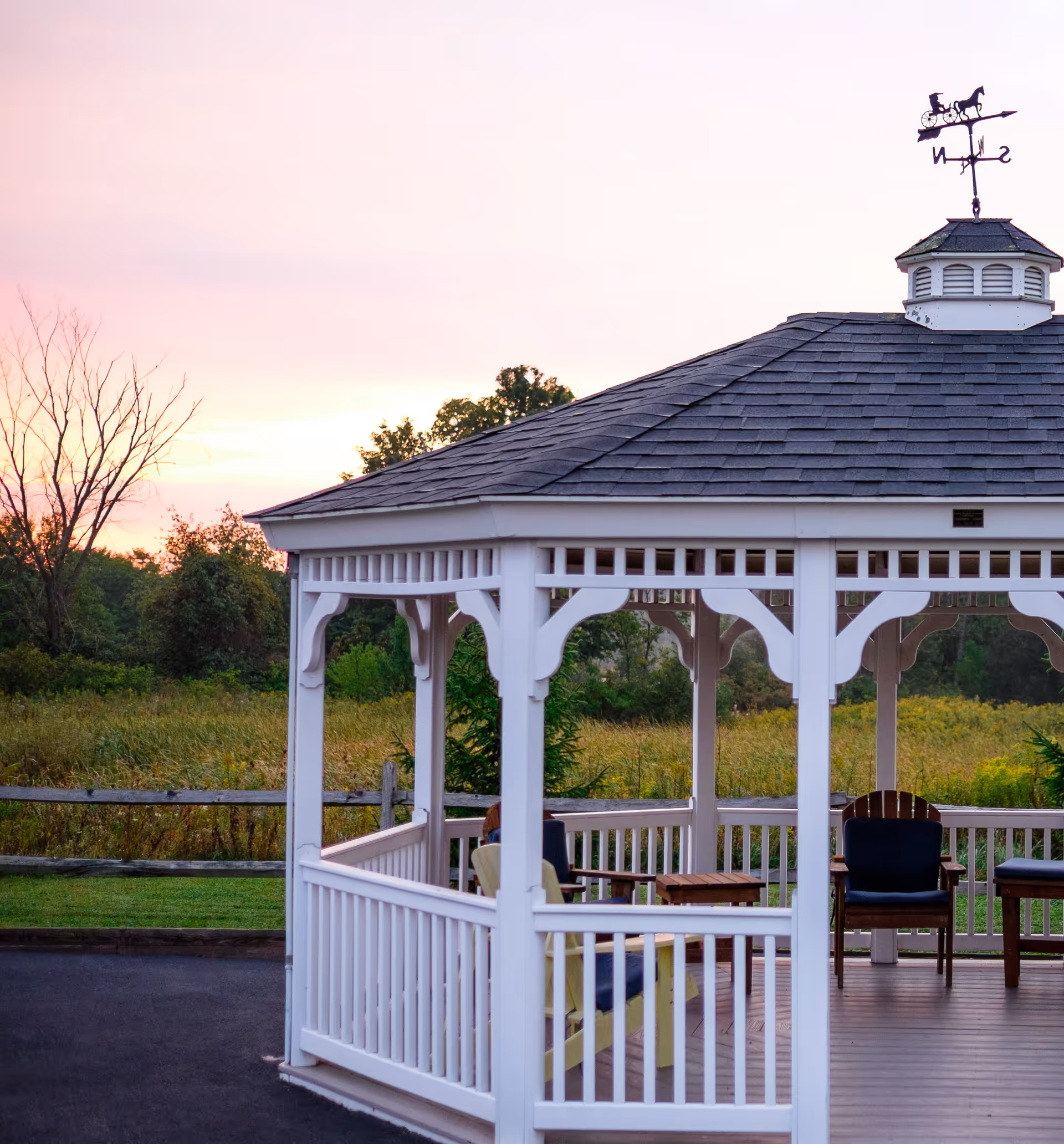 White wooden gazebo with a shingled roof and a weather vane on top, situated outdoors near a grassy field and trees during sunset. Inside the gazebo are several chairs and a small table.