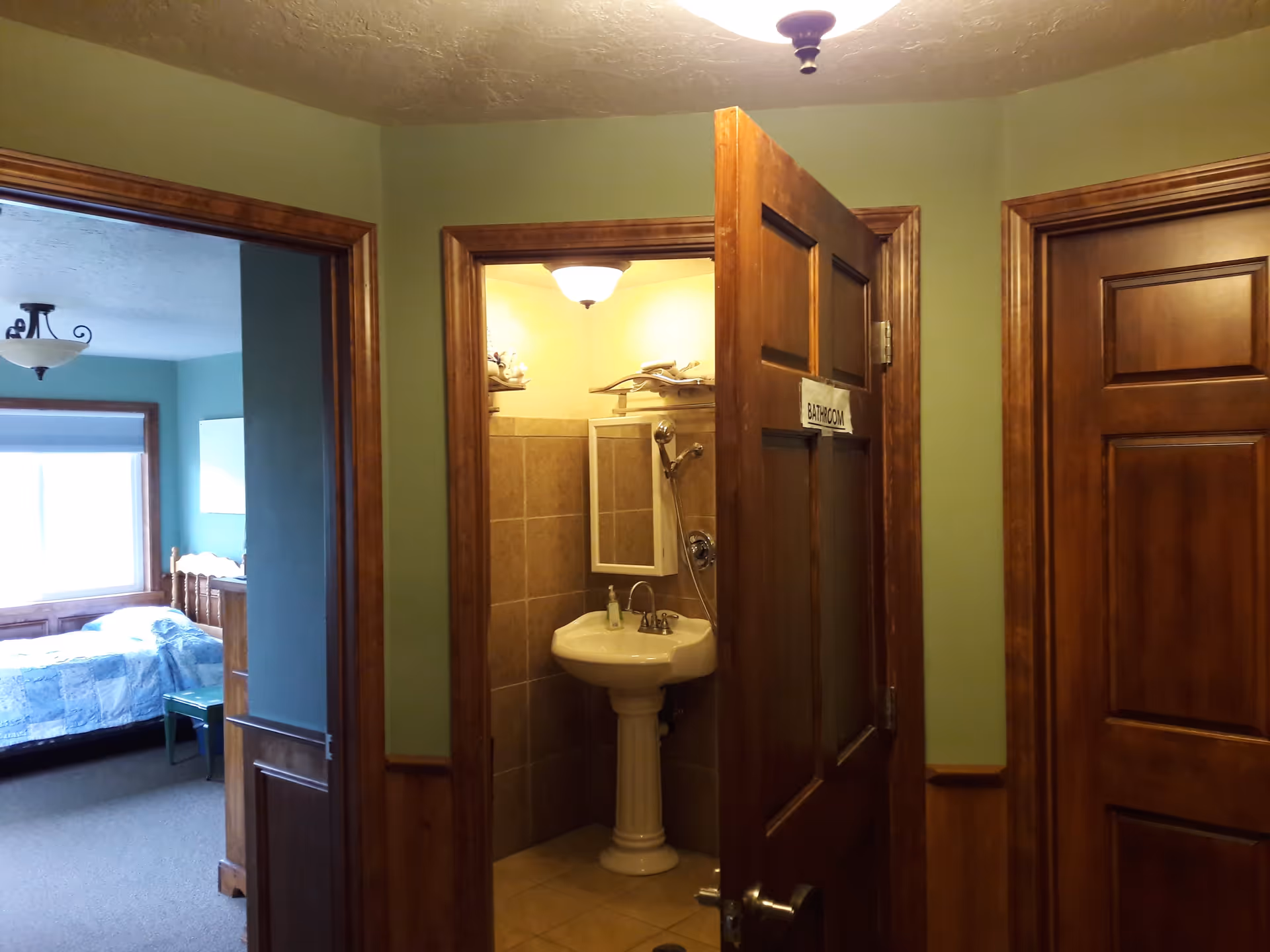 Hallway with an open door labeled "BATHROOM" revealing a pedestal sink and tiled shower, with a bedroom visible to the left.