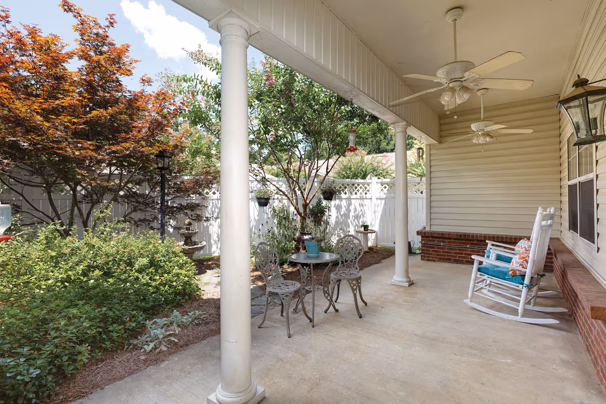 Covered outdoor patio area with white columns, two ceiling fans, a small round metal table with two matching chairs, and two white rocking chairs with cushions. The patio overlooks a garden with various green plants and a tree with red leaves, enclosed by a white fence.