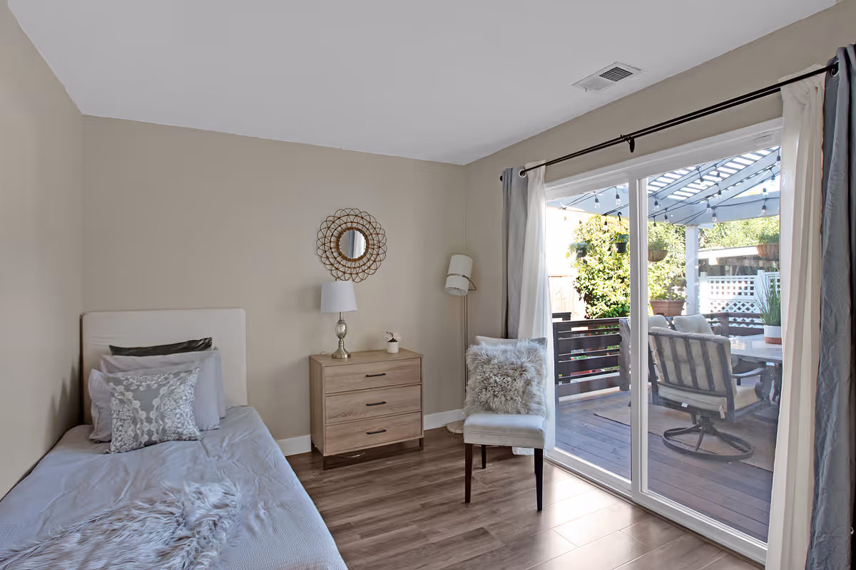 A cozy bedroom with a single bed adorned with multiple pillows and a soft throw blanket. Next to the bed is a wooden nightstand with a lamp and a small decorative item. A decorative round mirror hangs on the beige wall above the nightstand. A cushioned chair with a fluffy pillow is placed near a large sliding glass door that opens to an outdoor patio area with seating and greenery.