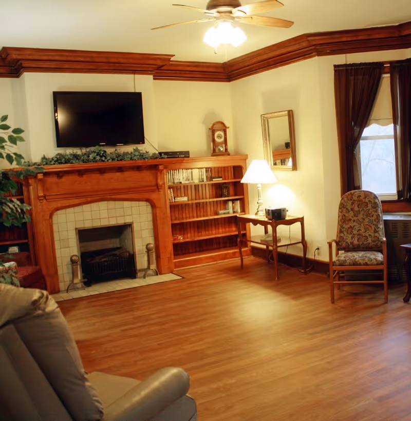 A cozy living room with wooden flooring, a wooden fireplace with a mounted flat-screen TV above it, a bookshelf filled with books, a small wooden side table with a lamp and a radio, a floral upholstered armchair, and windows with dark curtains.