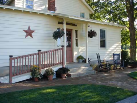 White house-style senior facility exterior with a porch, rocking chairs, potted plants, and star decor.