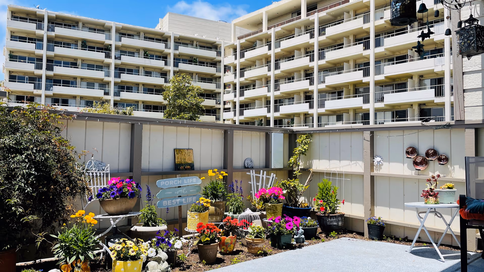 A bright outdoor patio area with various colorful potted flowers and plants arranged along a white fence. There is a white metal chair with a flower pot on it, a small white table with a plant, and decorative items including a sign that reads 'PORCH LIFE is the BEST LIFE'. In the background, a multi-story residential building with balconies is visible under a clear blue sky.
