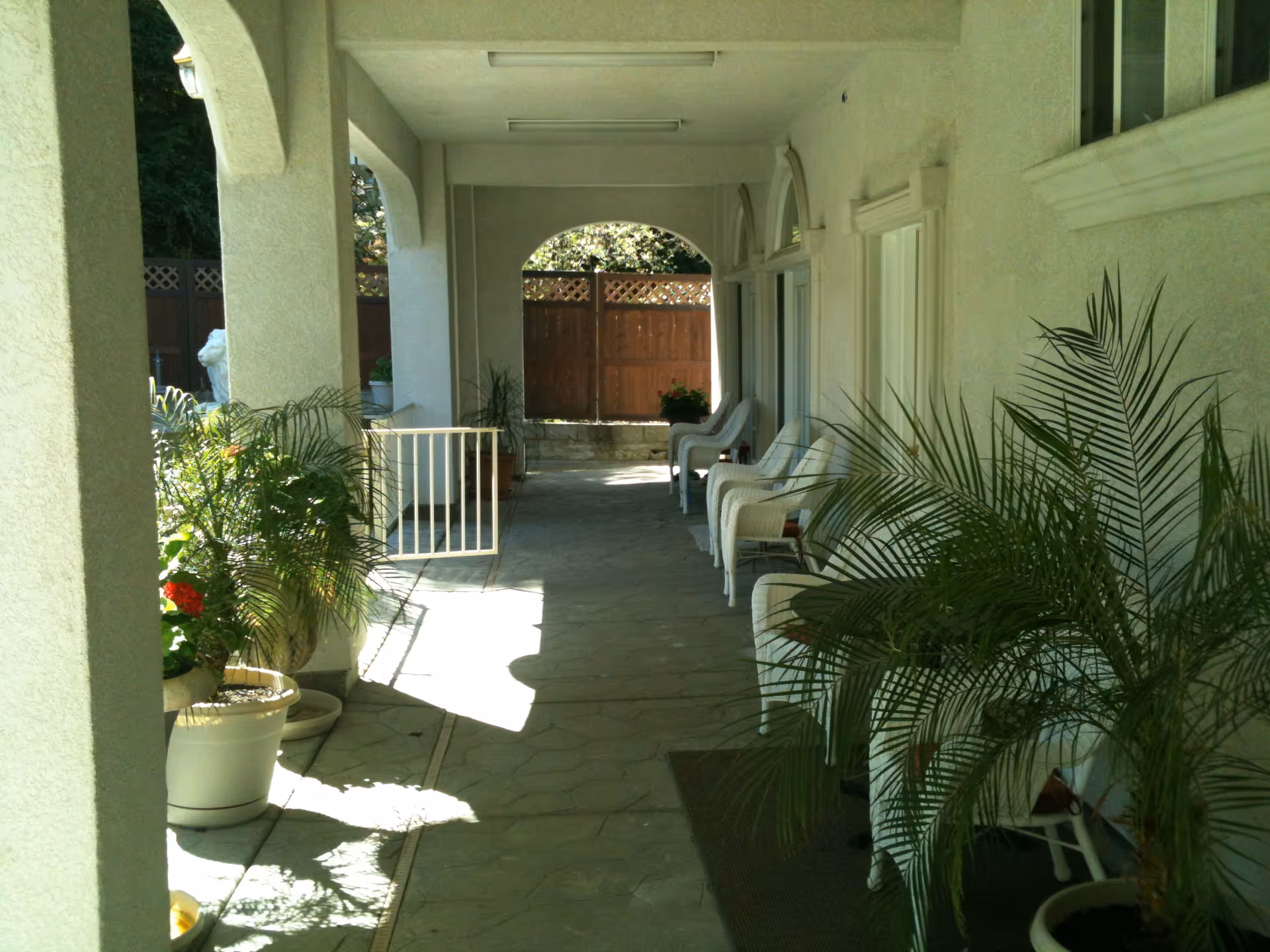 A covered outdoor patio area with white columns and arches, several white wicker chairs lined up against the wall, and multiple potted plants along the walkway. A wooden fence is visible at the end of the patio.