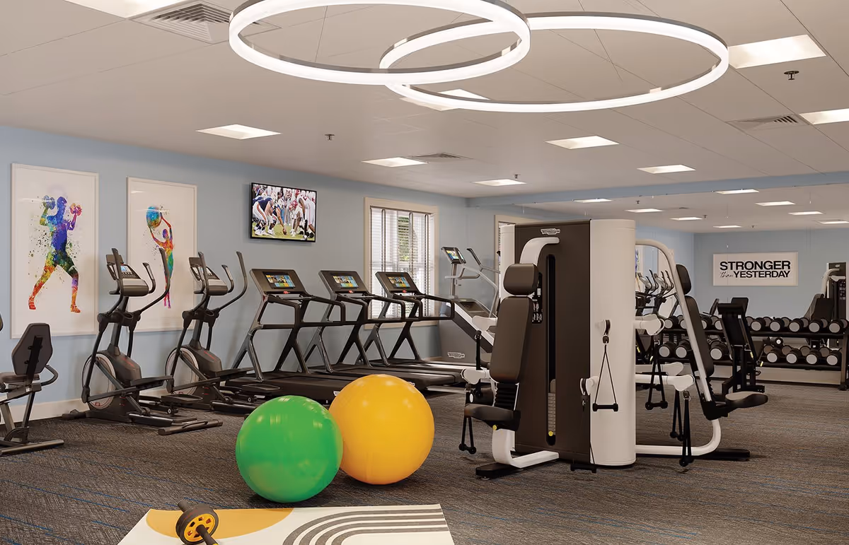 A modern fitness room with exercise equipment including treadmills, elliptical machines, a multi-functional weight machine, and free weights. Two large exercise balls, one green and one yellow, are on the floor. The walls are light blue with colorful sports-themed artwork and a motivational sign that reads 'STRONGER than YESTERDAY'. A TV is mounted on the wall showing a football game.
