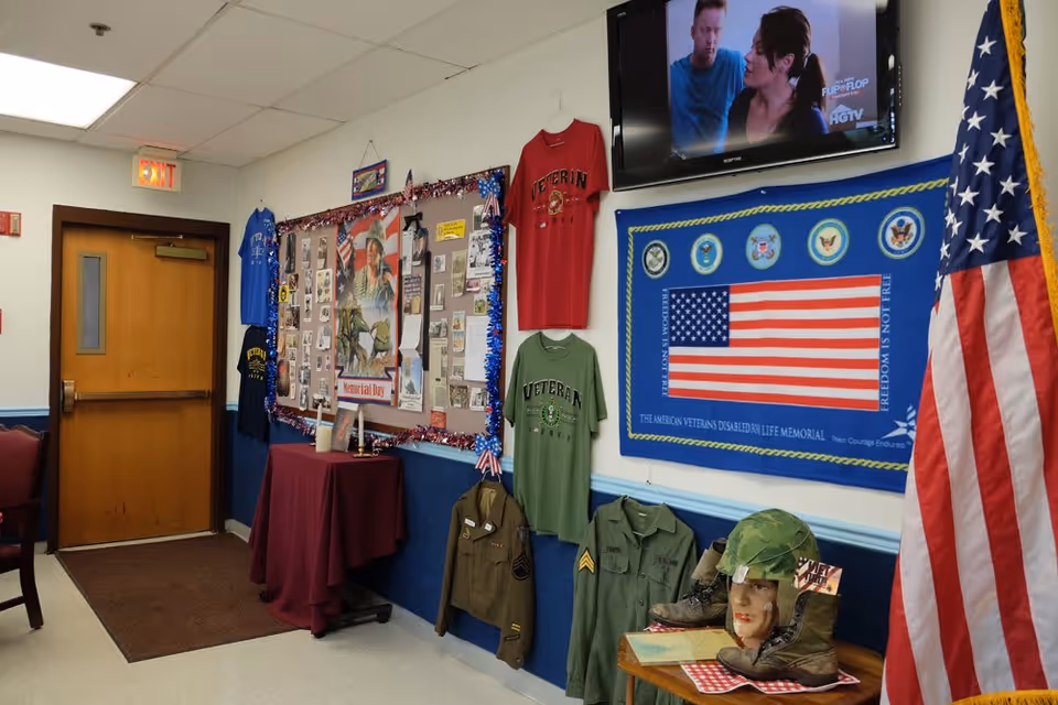 Interior hallway of a senior living facility decorated with military and veteran memorabilia including shirts, jackets, a flag, boots, and a mannequin head with a helmet. A bulletin board with photos and notes is displayed on the wall, and a television is mounted above showing a program. An American flag stands in the corner near a wooden door with an exit sign above it.