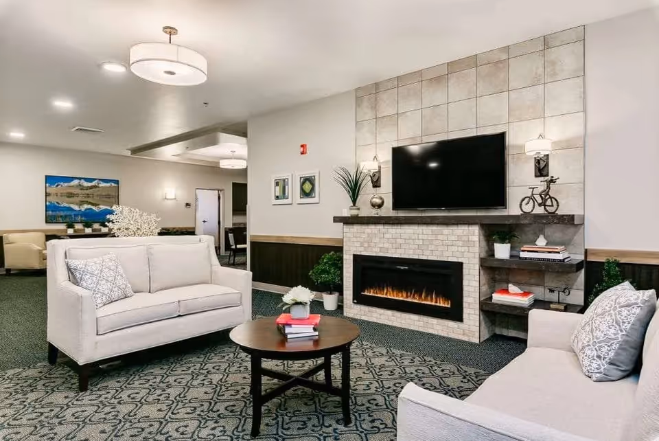 A cozy senior living facility common area with two beige sofas facing a modern electric fireplace topped with a flat-screen TV. The room features a patterned carpet, a round wooden coffee table with books and a small plant, wall art, and soft lighting fixtures on the ceiling and walls.