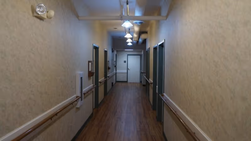 Long, well-lit interior corridor with wood flooring, handrails and doors on both sides in an assisted living residence.
