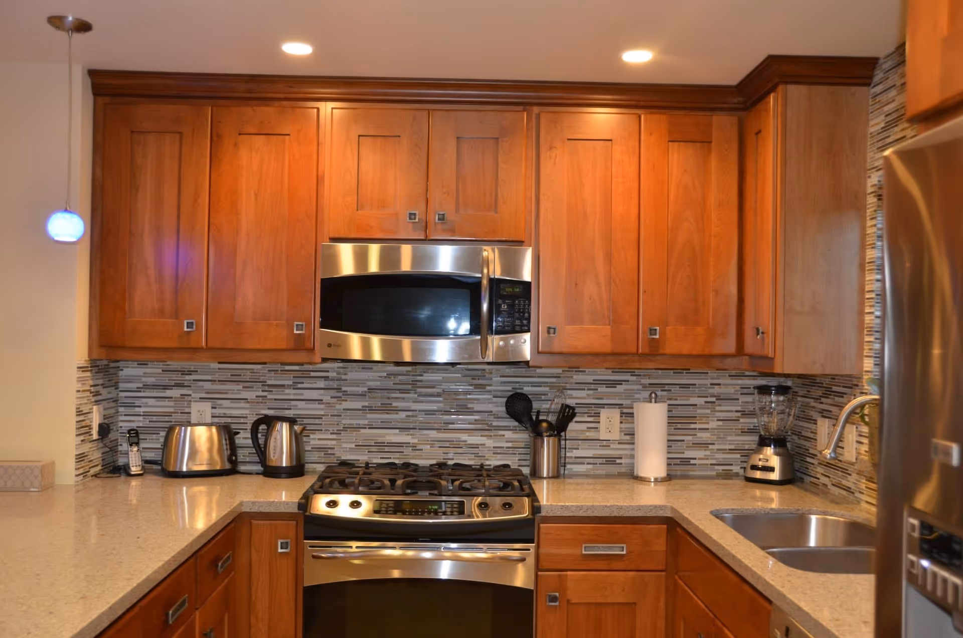 Modern kitchen with wooden cabinets, a stainless steel microwave above a gas stove, a toaster, electric kettle, blender, paper towel holder, and a sink with a faucet. The backsplash features a mosaic tile design in neutral colors.