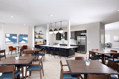 Bright communal dining room with multiple wooden tables and chairs and an open kitchen island with pendant lights.