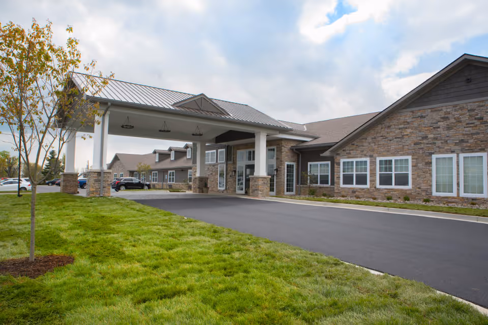 Exterior view of Boonesboro Trail Senior Living facility showing a single-story building with stone and siding facade, a covered entrance with white pillars, a paved driveway, green lawn, and a cloudy sky.