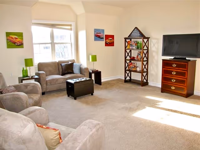 A bright living room with beige carpet and light-colored walls. The room features a gray loveseat with two pillows, two matching armchairs, a black ottoman in the center, and two small side tables with green lamps and decorative bottles. On the right side, there is a wooden chest of drawers with a flat-screen TV on top and a wooden shelf unit with various items. Three colorful car-themed paintings hang on the walls, and sunlight streams in through a large window.