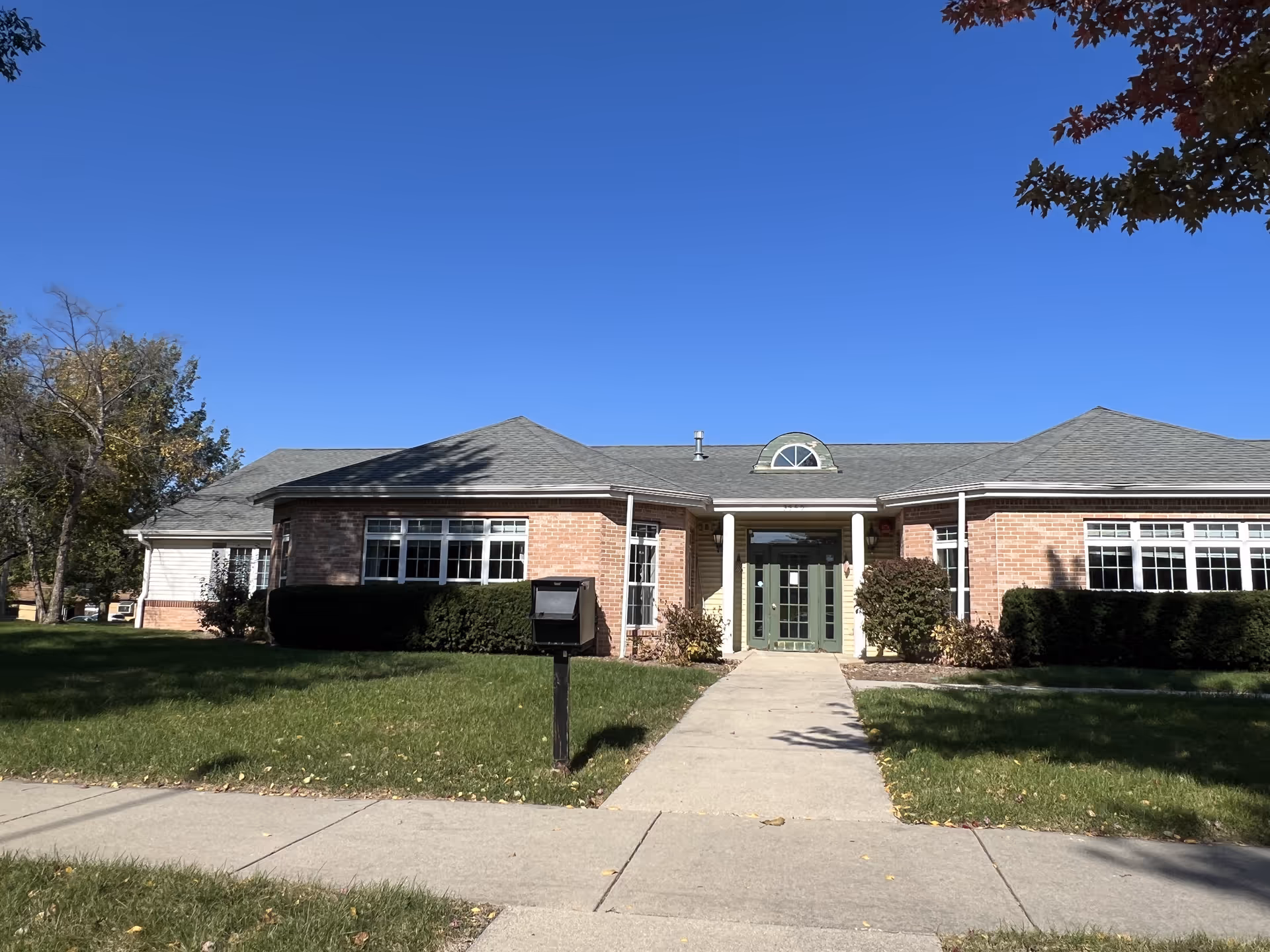 Front exterior view of a single-story brick building with a gray roof, green double doors, and large windows. There is a concrete walkway leading to the entrance, surrounded by green grass and bushes, under a clear blue sky.