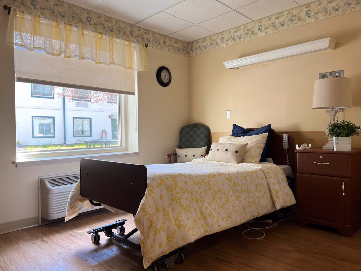 A single bed with yellow floral bedding in a well-lit room. The bed has a dark wooden headboard and footboard, and there are two pillows and a cushion on it. Next to the bed is a wooden nightstand with a lamp and a small potted plant. A green cushioned chair is placed in the corner near the bed. A large window with a yellow valance lets in natural light, showing an exterior view of another building. The room has light-colored walls with a floral border near the ceiling and a wooden floor.