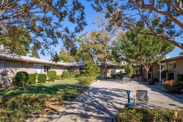 Outdoor courtyard area of Southland Care Center with a paved walkway, green grass, bushes, and trees under a clear blue sky. Single-story buildings surround the courtyard.