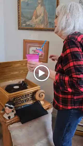An elderly woman with white hair wearing a red and black plaid shirt stands next to a vintage record player. She holds a pink cup with a blue straw and a decorative umbrella. Behind her on the wall are two framed pictures, one depicting a religious scene and the other a smaller artwork. A small figurine of a person wearing a hat is placed near the record player.