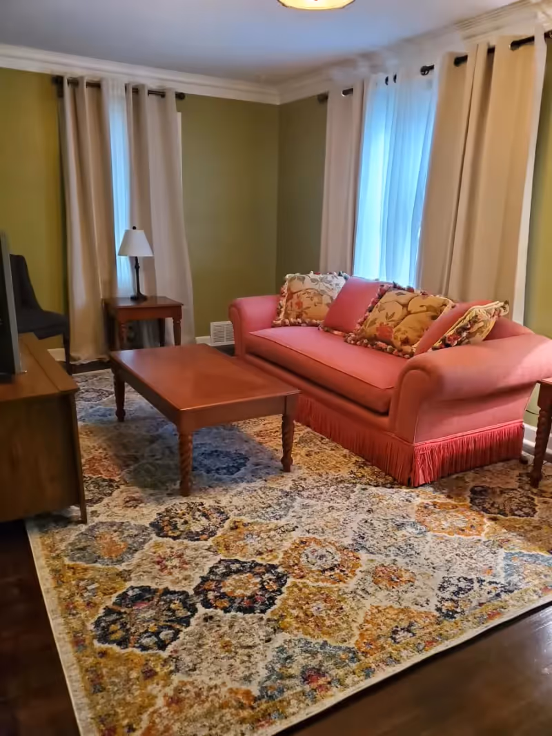 A cozy living room with olive green walls, two windows with beige curtains, a pink sofa with floral patterned cushions, a wooden coffee table, a side table with a lamp, and a colorful patterned area rug covering the wooden floor.