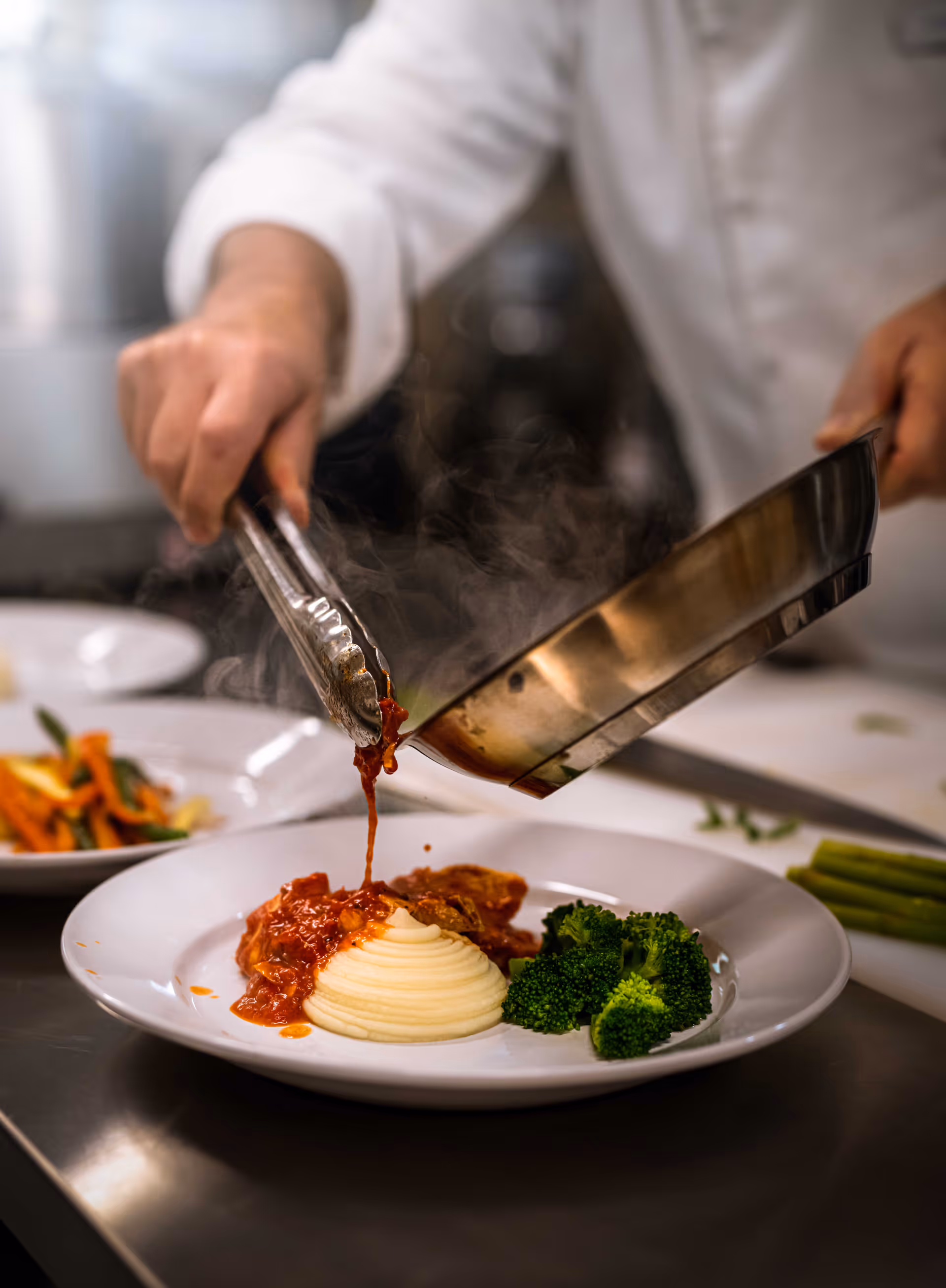 A chef in a white uniform uses tongs to pour steaming tomato sauce from a pan onto a plate with mashed potatoes and broccoli in a kitchen setting.