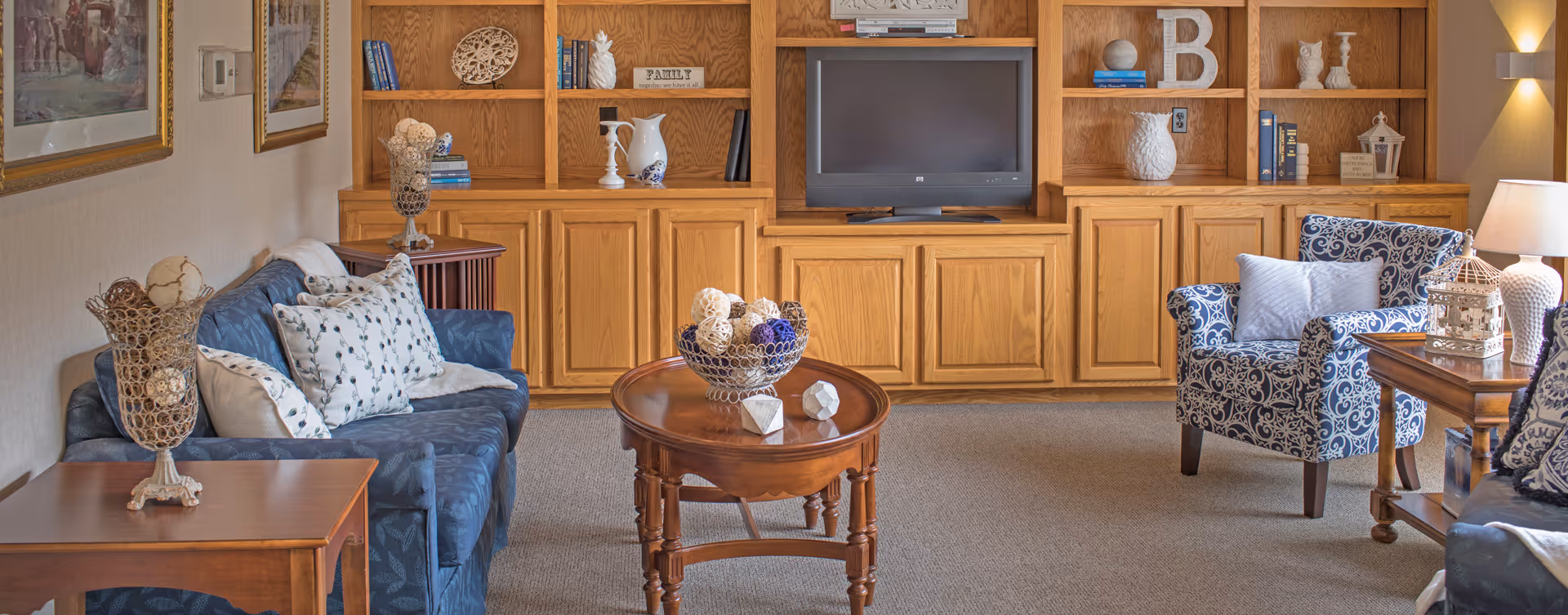 A cozy living room with a blue patterned sofa and armchair, wooden coffee and side tables, and a large wooden built-in shelving unit with a television in the center. The shelves are decorated with books, vases, and decorative items. The room is carpeted and softly lit by a table lamp.