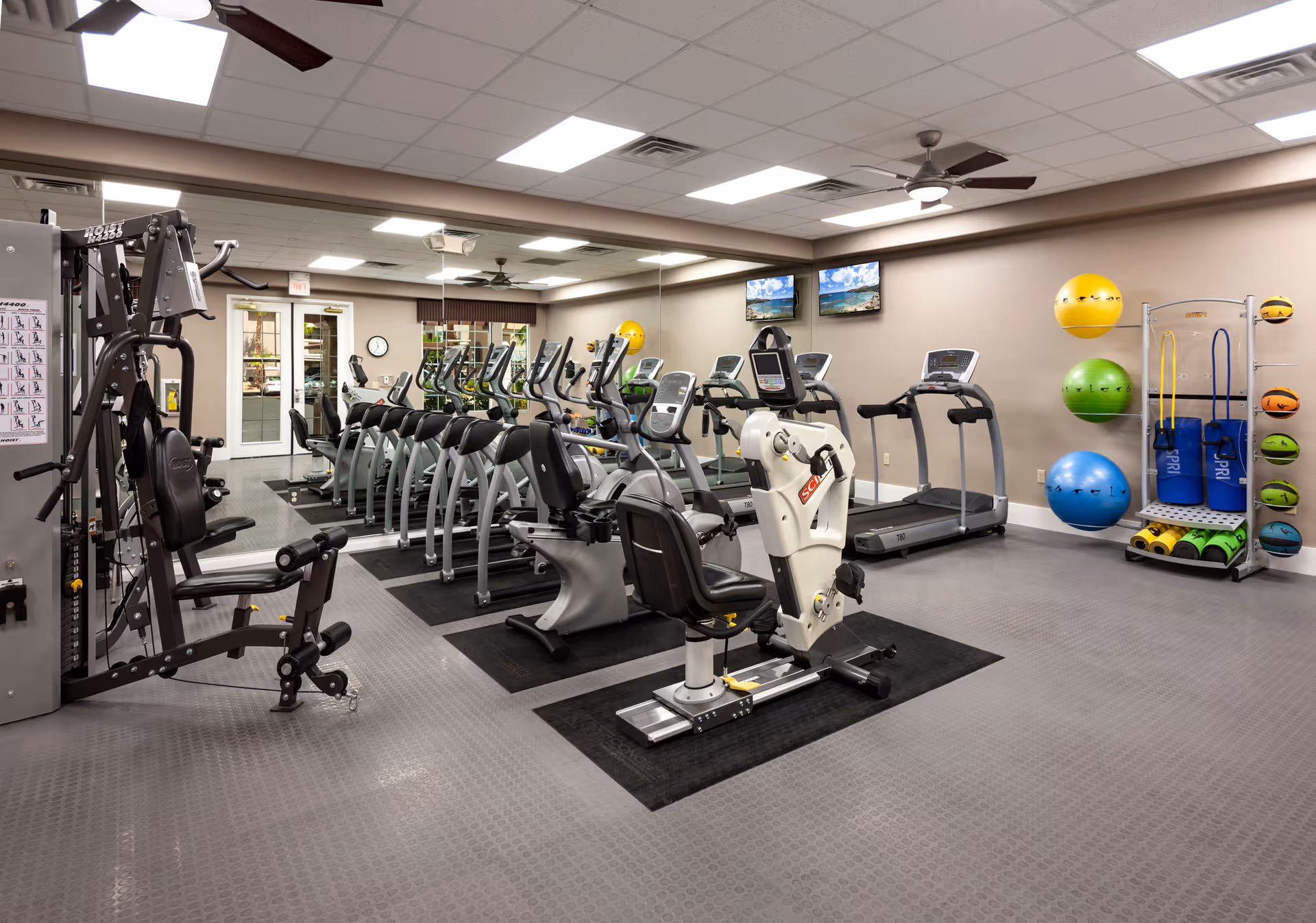 A well-lit fitness room with various exercise equipment including treadmills, stationary bikes, a multi-function weight machine, and a rack with colorful exercise balls and resistance bands. The room has a large mirror on one wall, two ceiling fans, and two wall-mounted TVs displaying a beach scene.
