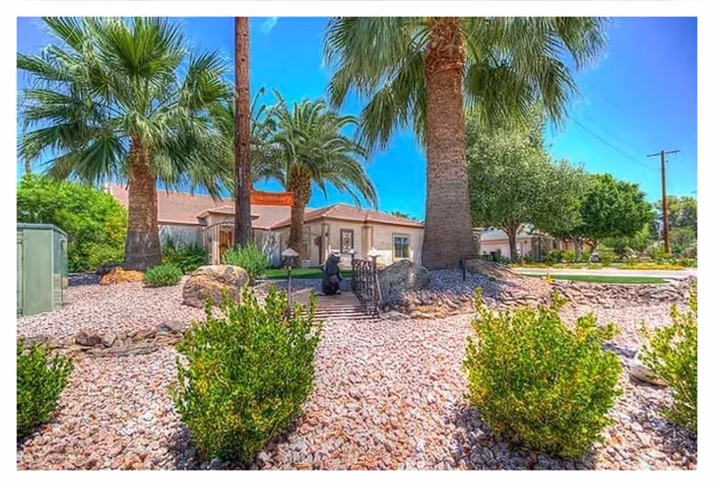 Outdoor view of a residential facility with palm trees, bushes, rocks, and a clear blue sky. The building has a light-colored exterior with a red-tiled roof and a small staircase leading to the entrance.