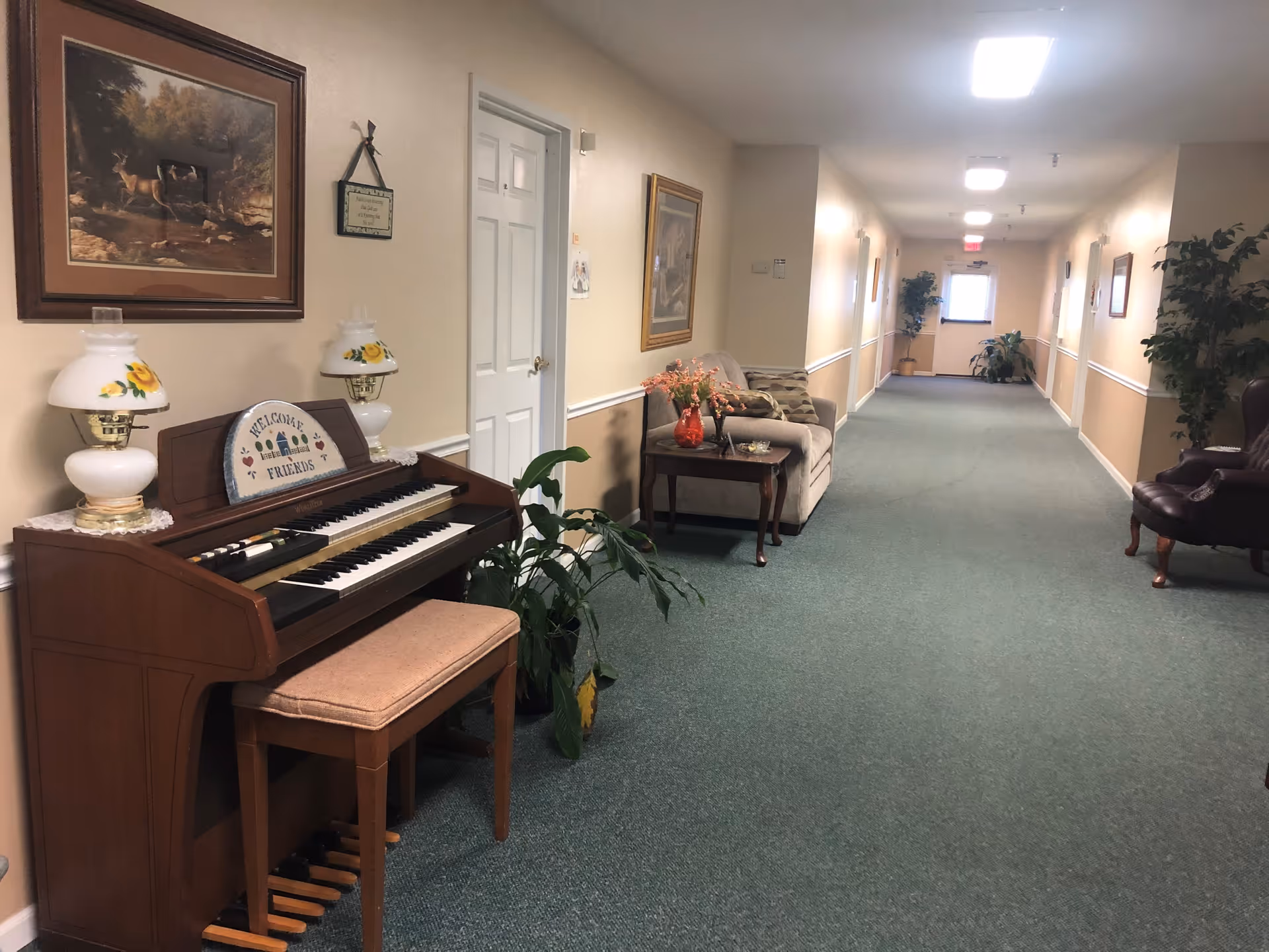 A long, carpeted hallway in a senior living facility with beige walls and white trim. On the left side, there is a wooden organ with two decorative lamps and a sign that says 'Welcome Friends.' Above the organ is a framed painting of deer in a forest. Further down the hallway, there is a beige couch with patterned pillows and a small wooden side table holding a red vase with flowers. Several potted plants are placed along the hallway, and framed pictures hang on the walls. The hallway is well-lit with ceiling lights and ends with a door and window letting in natural light.