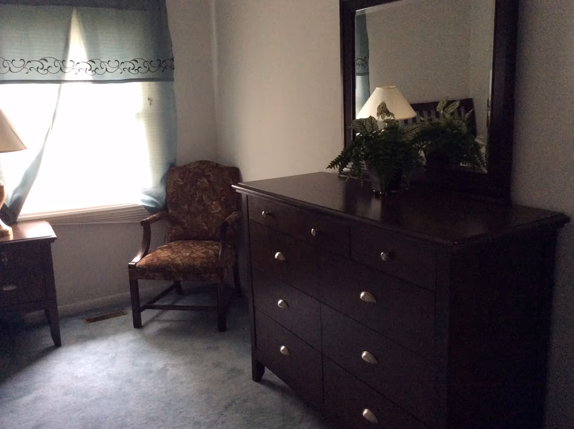 A dimly lit bedroom corner featuring a dark wooden dresser with a large mirror, a potted fern on top, a floral upholstered armchair, a small wooden side table with a lamp, and a window with blue curtains partially drawn.
