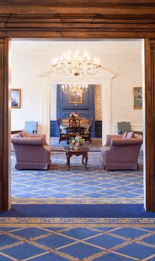 View through a wooden doorway into a formal sitting area with two upholstered armchairs and a small wooden table with a flower arrangement. Beyond this room is a dining area with a wooden dining table, chairs, and a chandelier hanging from the ceiling. The rooms have patterned blue carpeting and light-colored walls with framed artwork.