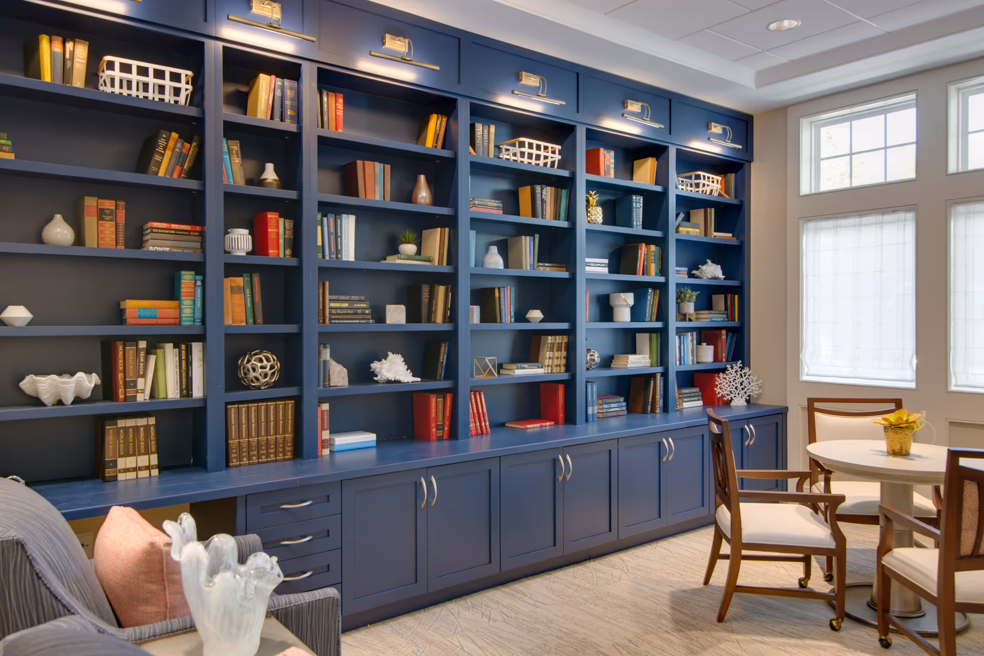 A cozy interior room featuring a large blue built-in bookshelf filled with books and decorative items. To the right, there is a round table with four wooden chairs with white cushions. The room is well-lit by large windows with white blinds, and a comfortable armchair with a pink pillow is partially visible on the left.
